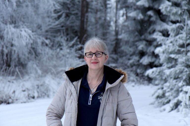 Harmaatukkainen nainen seisoo ulkona talvisen metsän laidalla